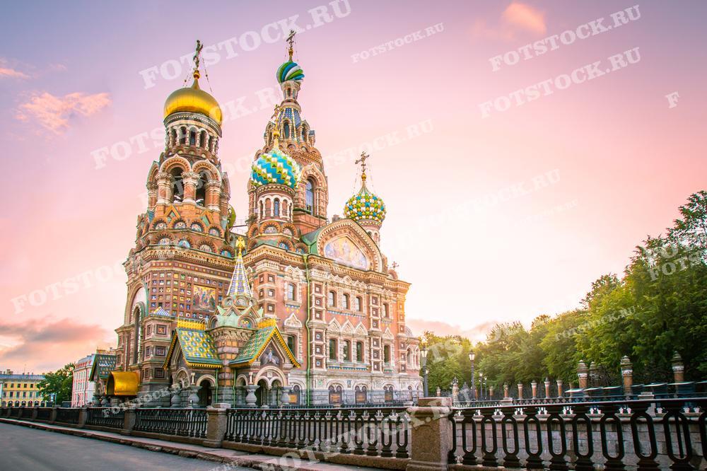 На фотографии изображен Храм Спаса на Крови в Санкт-Петербурге на фоне заката. Видна набережная канала Грибоедова и окружающие деревья. Фото может использоваться для туристических буклетов, открыток и рекламных материалов.